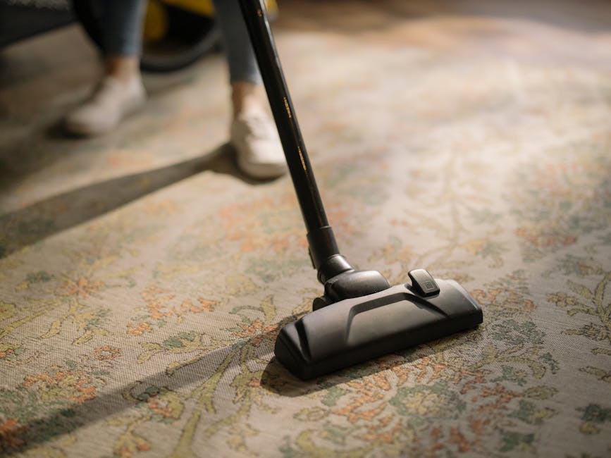 A close-up view of a vacuum cleaner head being used to clean an ornate, patterned area rug with floral motifs in shades of beige, green, and orange. The vacuum cleaner, operated by an individual weari