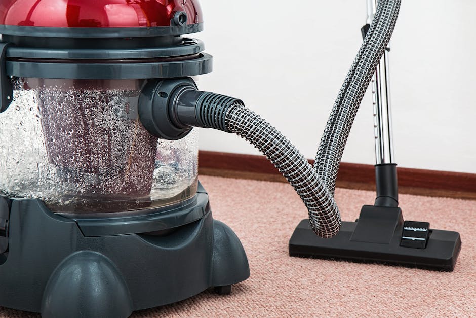 A close-up view of a vacuum cleaner head being used to clean an ornate, patterned area rug with floral motifs in shades of beige, green, and orange. The vacuum cleaner, operated by an individual weari