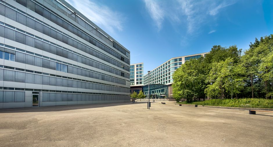 An exterior view of Stockley Park office buildings featuring modern, glass-fronted facades with metallic and concrete surfaces. The paved open area in the foreground appears clean and well-maintained,