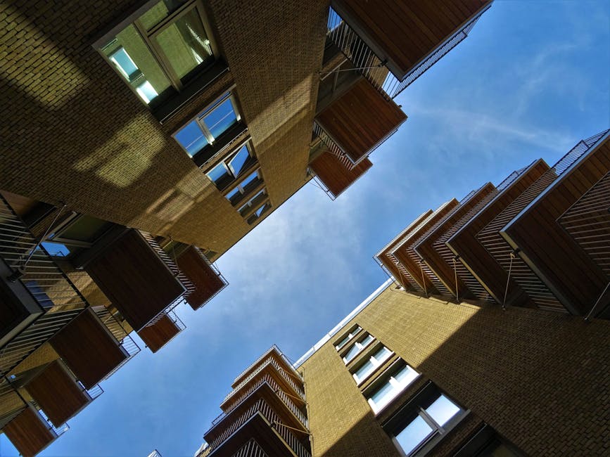 A view looking upward at the facades of modern residential apartment buildings in Uxbridge, displaying brick exteriors, multiple balconies with metal railings, and large glass windows. The bright blue